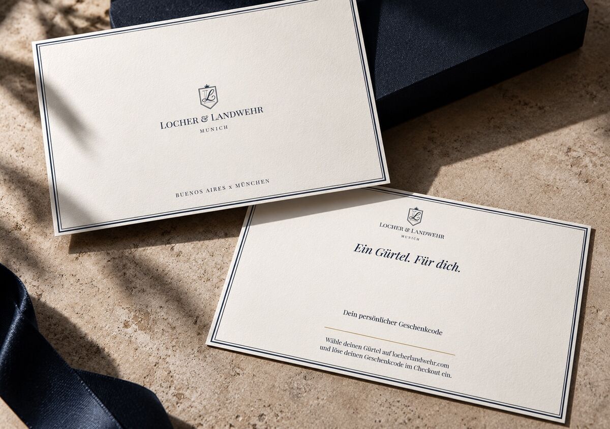 Die beiden gedruckten Karten neben der navyfarbenen Geschenkbox: vorn das Wappen, hinten &bdquo;Ein Gürtel. Für dich.&ldquo; und die Linie für den Geschenkcode.